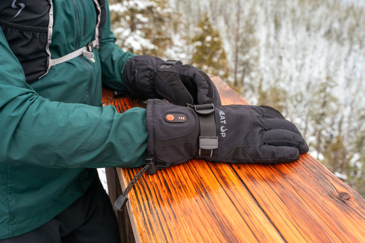 Person in a green jacket wearing Heat Up Mountain heated gloves, hands on a wooden railing in winter