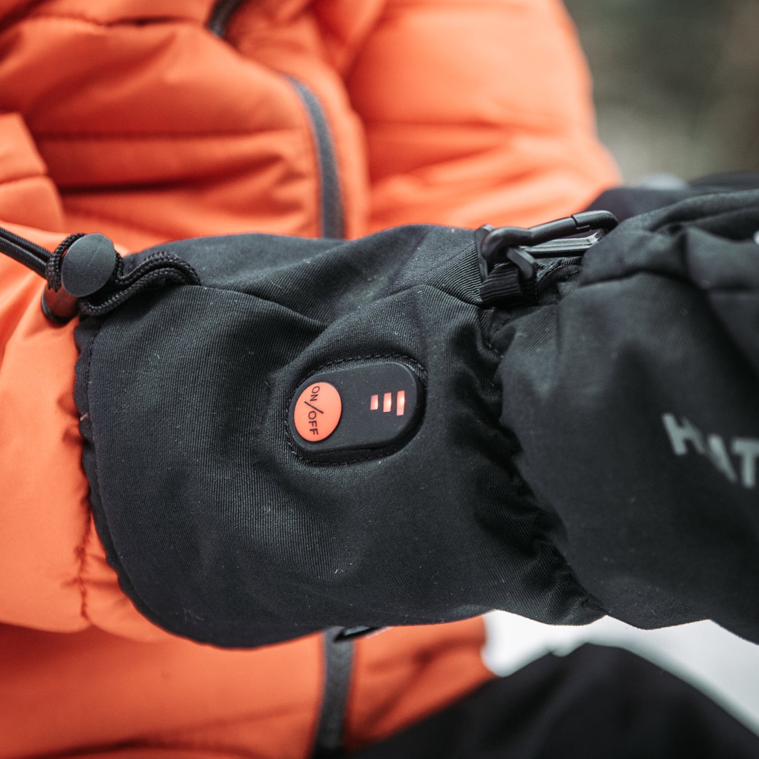 Black heated glove with visible brand logo, worn by a person in an orange jacket.