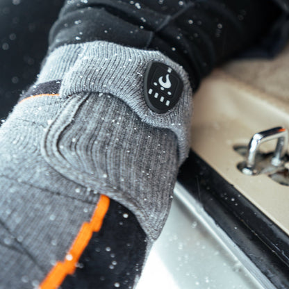 Tige d'un bas chauffant Merino Wool+ Heat Up avec batterie et bouton de chaleur, près d'une botte de ski dans la neige