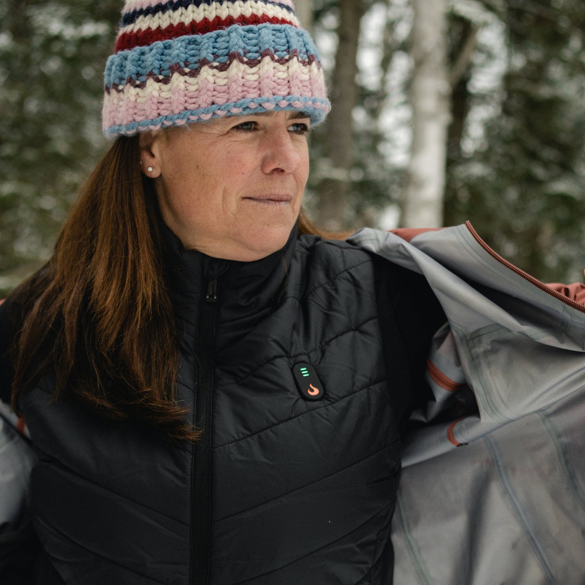 Woman in a knit hat opening her jacket to show the Heat Up women's heated vest underneath, heat button lit
